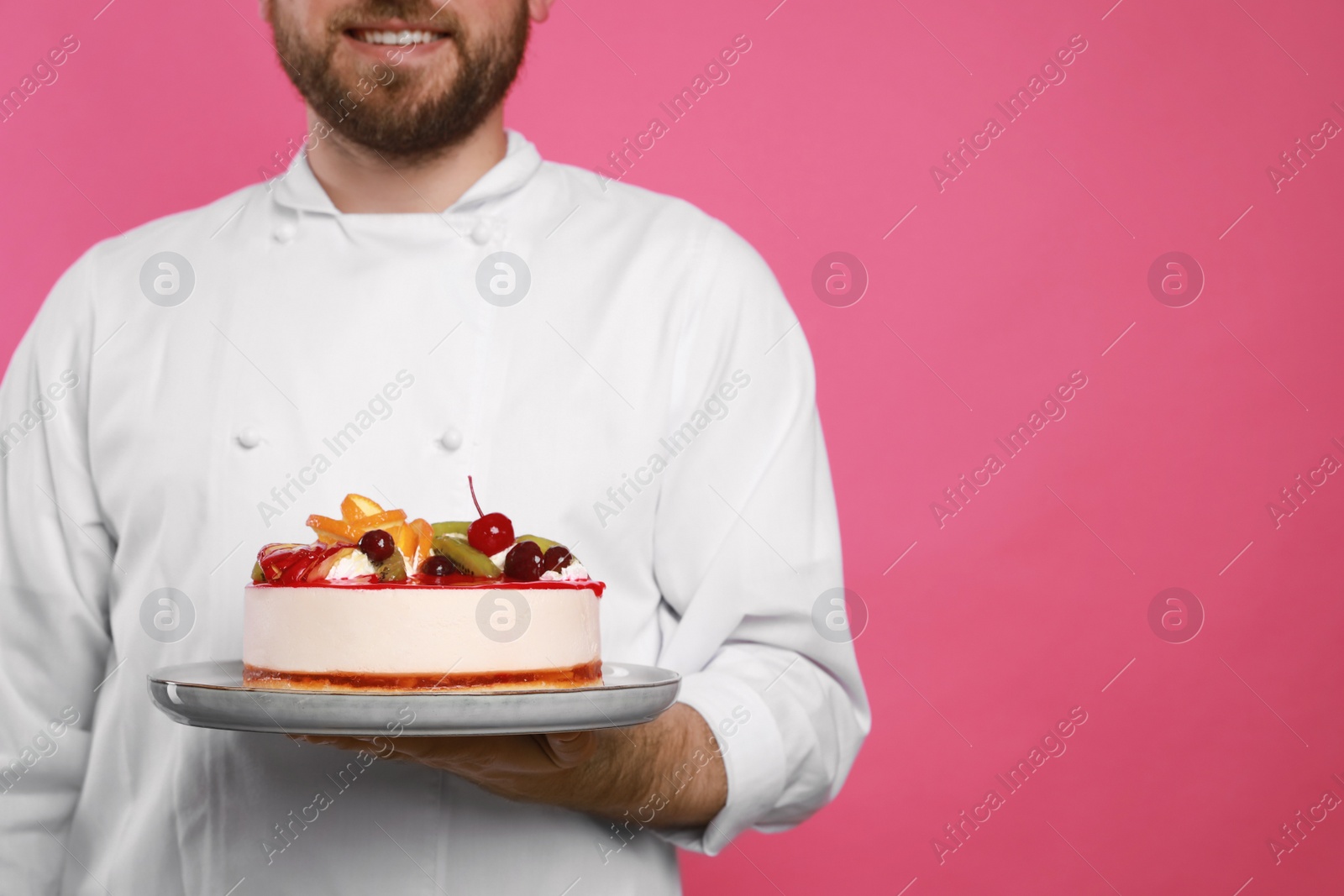 Photo of Happy professional confectioner in uniform holding delicious cake on pink background, closeup. Space for text