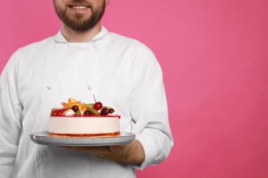 Happy professional confectioner in uniform holding delicious cake on pink background, closeup. Space for text