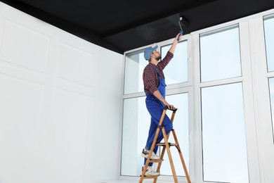 Photo of Worker in uniform painting ceiling with roller on stepladder indoors