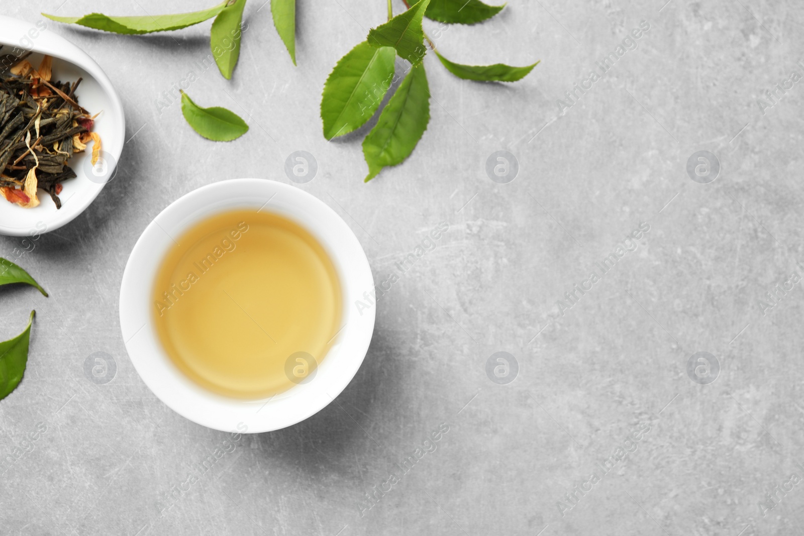 Photo of Cup of green tea, dry and fresh leaves on grey table, flat lay. Space for text