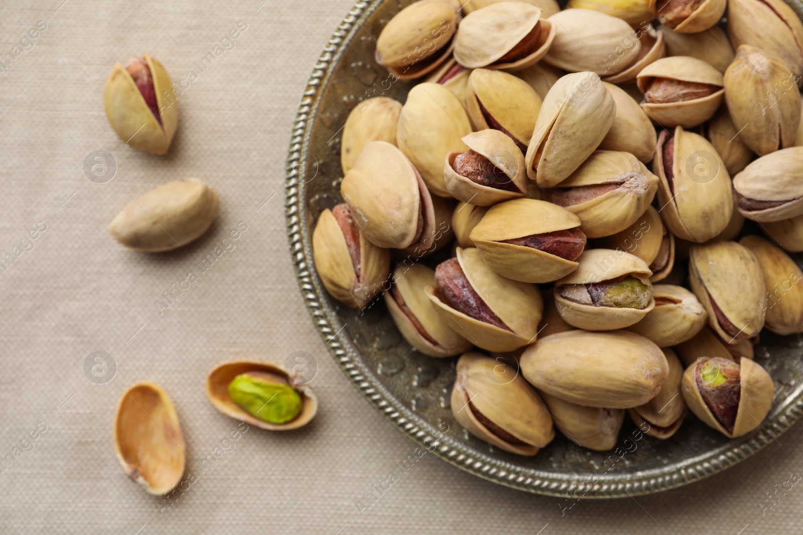 Photo of Plate and pistachio nuts on beige tablecloth, flat lay