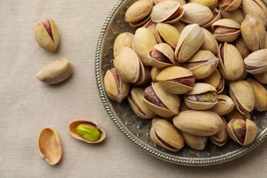 Photo of Plate and pistachio nuts on beige tablecloth, flat lay