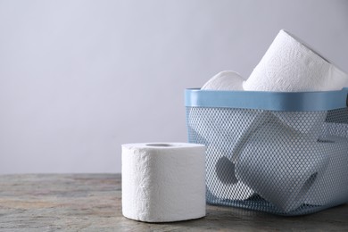 Photo of Toilet paper rolls in basket on textured table near light grey wall, space for text