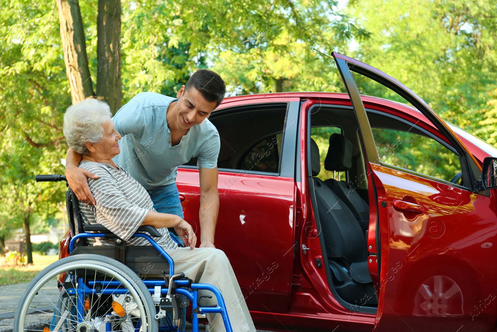 Photo of Young man helping disabled senior woman in wheelchair to get into car outdoors