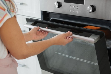 Photo of Young woman opening electric oven in kitchen, closeup