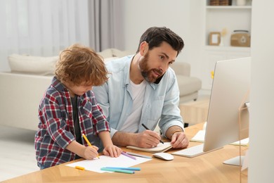 Photo of Man working remotely at home. Busy father talking on smartphone while his son drawing at desk