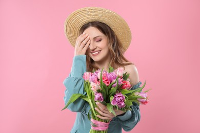 Happy young woman in straw hat holding bouquet of beautiful tulips on pink background