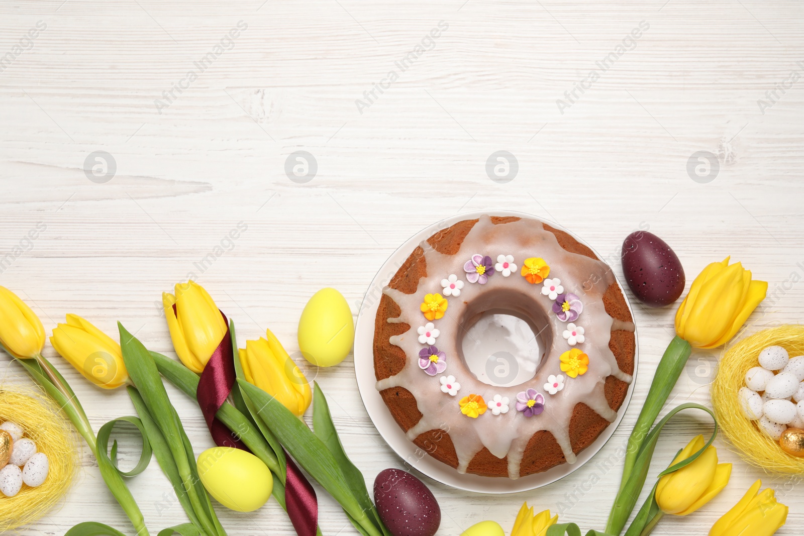Photo of Flat lay composition of delicious Easter cake decorated with sprinkles, eggs and beautiful tulips on white wooden table. Space for text