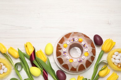 Flat lay composition of delicious Easter cake decorated with sprinkles, eggs and beautiful tulips on white wooden table. Space for text