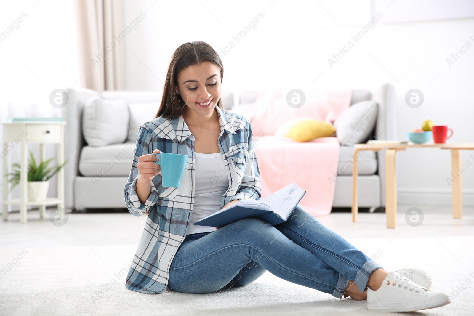 Photo of Beautiful young woman with cup of coffee reading book on floor at home