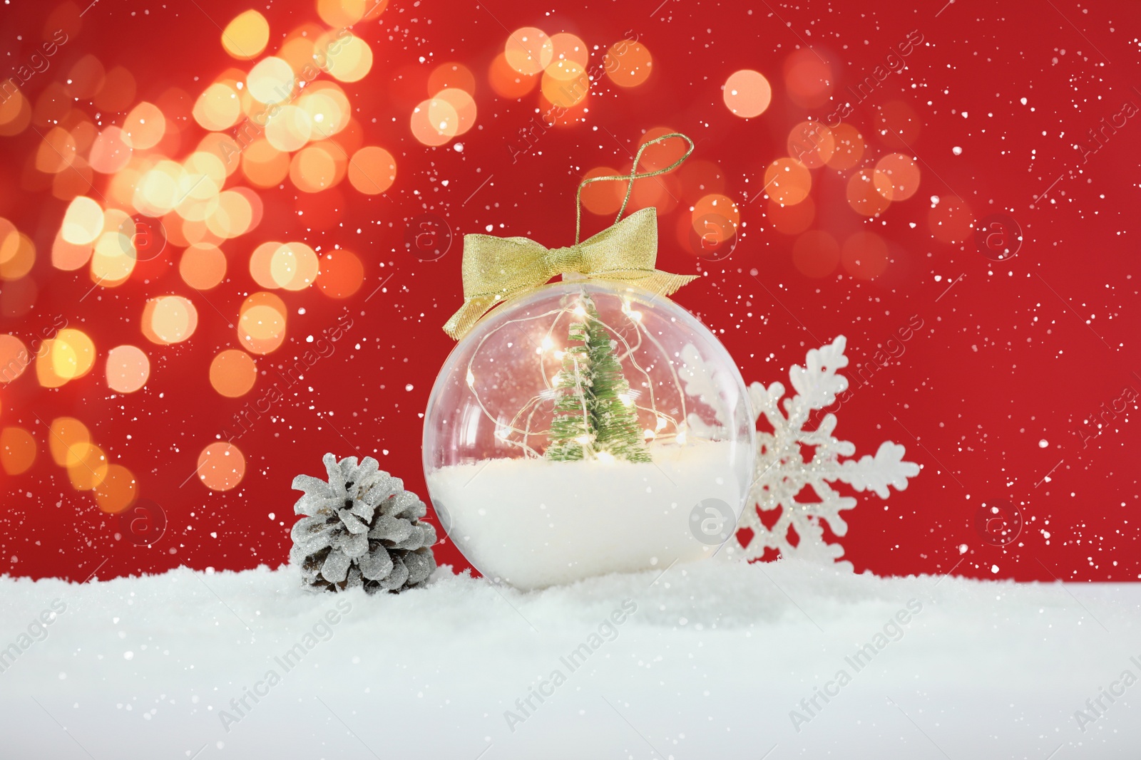 Image of Beautiful transparent Christmas ornament with small fir tree and fairy lights on snow against red background, bokeh effect