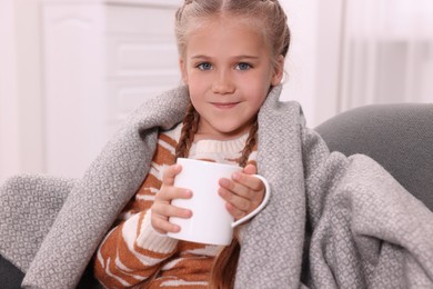 Happy girl under plaid with white ceramic mug on sofa at home
