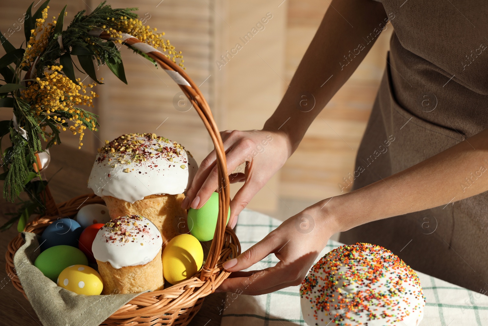 Photo of Woman putting dyed eggs in basket with traditional Easter cake at table, closeup