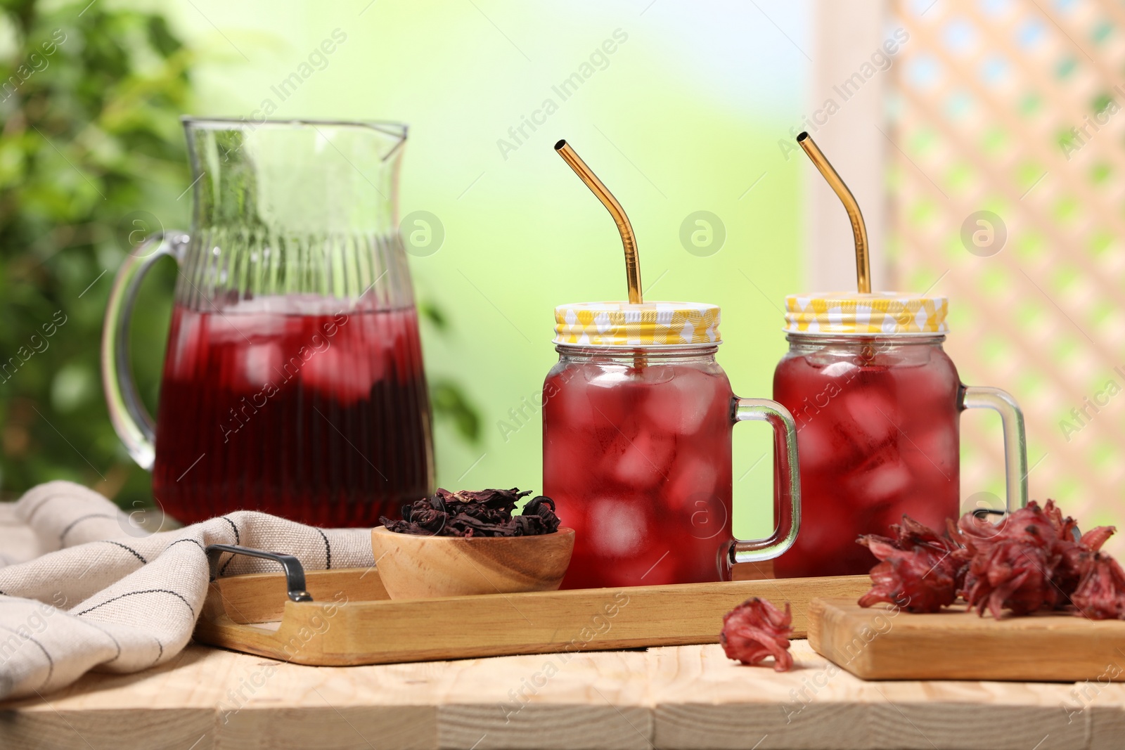 Photo of Refreshing hibiscus tea with ice cubes and roselle flowers on wooden table against blurred green background