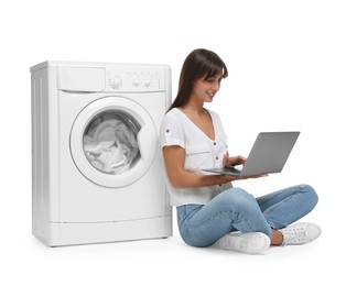 Photo of Beautiful woman using laptop near washing machine with laundry on white background