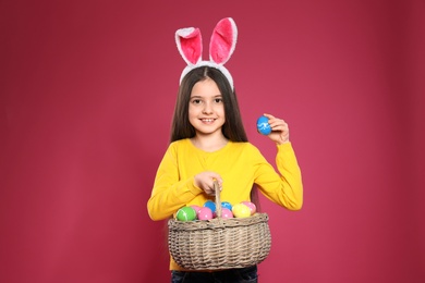 Little girl in bunny ears headband holding basket with Easter eggs on color background