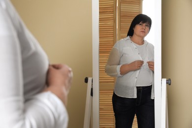Overweight woman trying to button up tight shirt in front of mirror at home