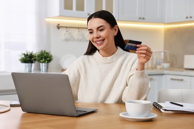 Photo of Happy young woman with credit card using laptop for shopping online at wooden table in kitchen