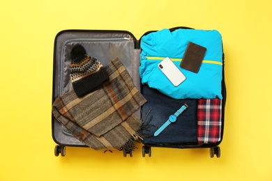 Open suitcase with warm clothes and accessories on color background, top view