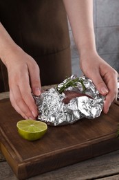 Woman wrapping meat in aluminum foil at wooden table, closeup