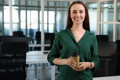Happy woman with paper cup of drink in office, space for text