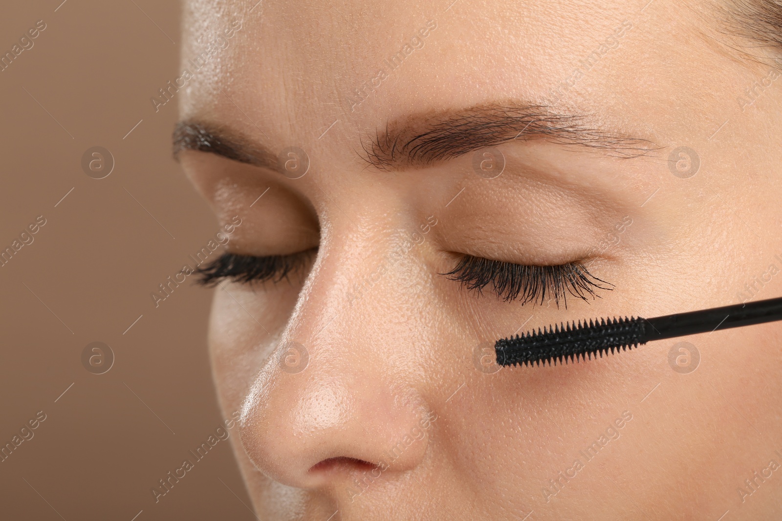 Photo of Woman applying mascara onto eyelashes against light brown background, closeup