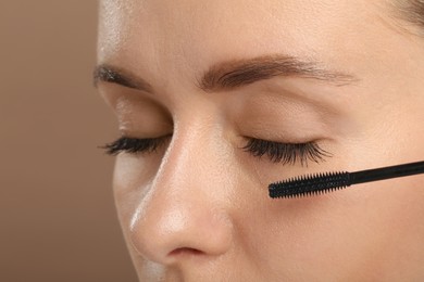 Woman applying mascara onto eyelashes against light brown background, closeup