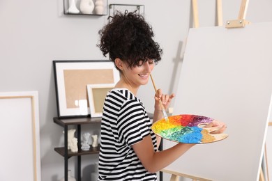 Photo of Young woman mixing paints on palette with brush near easel in studio