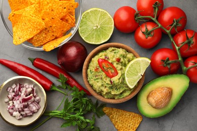 Photo of Bowl of delicious guacamole, nachos chips and ingredients on grey table, flat lay