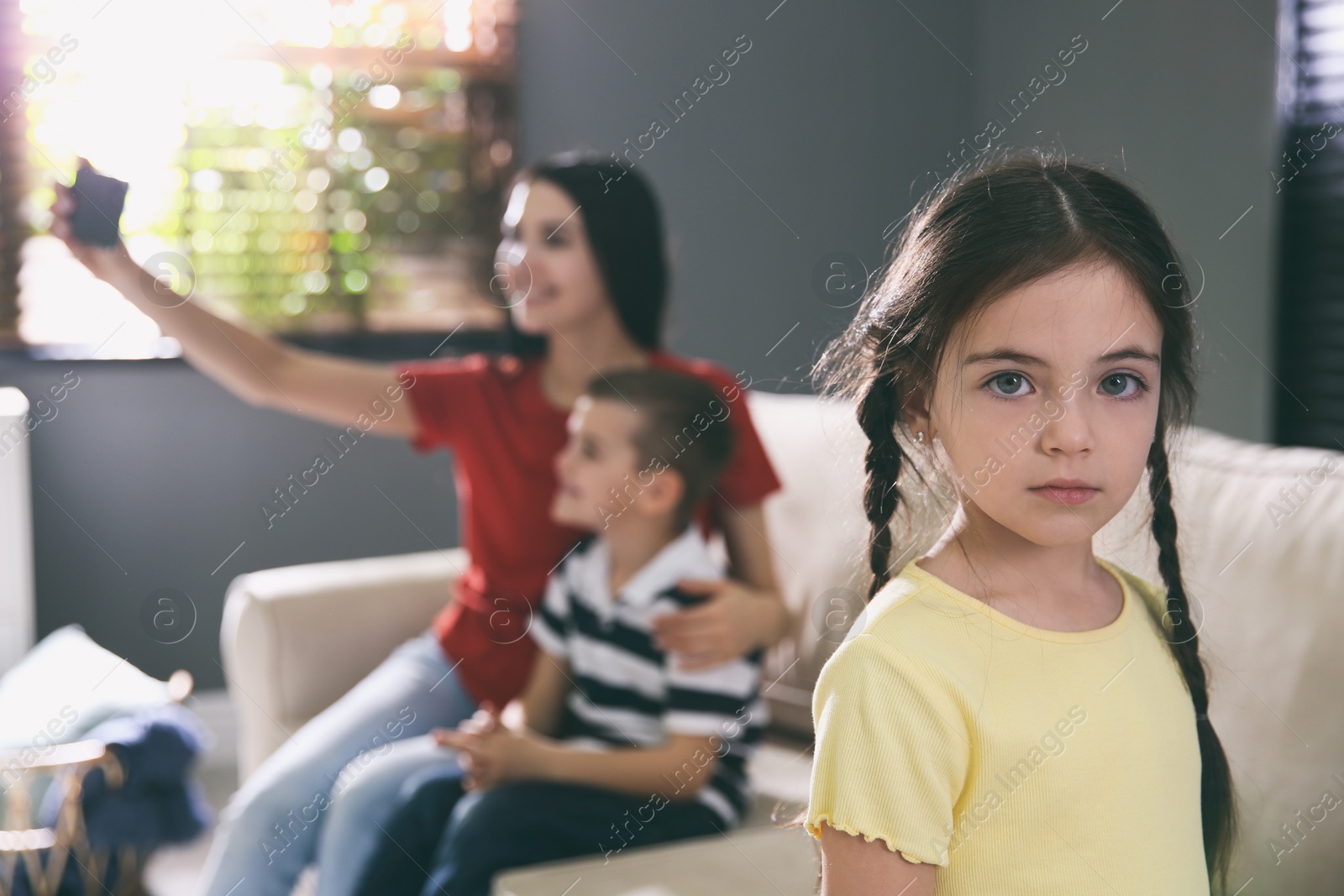 Photo of Unhappy little girl feeling jealous while mother taking selfie with her brother at home