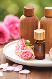 Bottles of rose essential oil and flowers on wooden table outdoors, closeup
