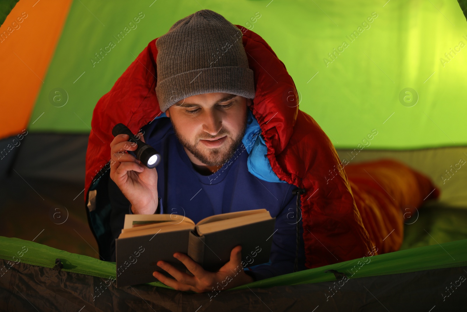 Photo of Young man with flashlight reading book in tent