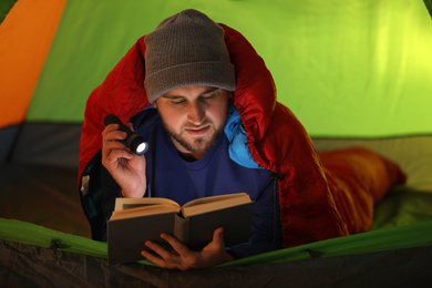 Young man with flashlight reading book in tent