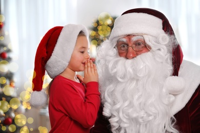 Photo of Little girl whispering in Santa Claus' ear near Christmas tree indoors