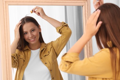 Beautiful woman applying serum onto hair near mirror indoors