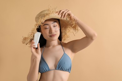 Photo of Beautiful young woman in straw hat with sunscreen on her face holding sun protection cream against beige background, space for text