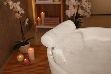 Photo of Tub with foamy water and soft bath pillow surrounded by candles indoors
