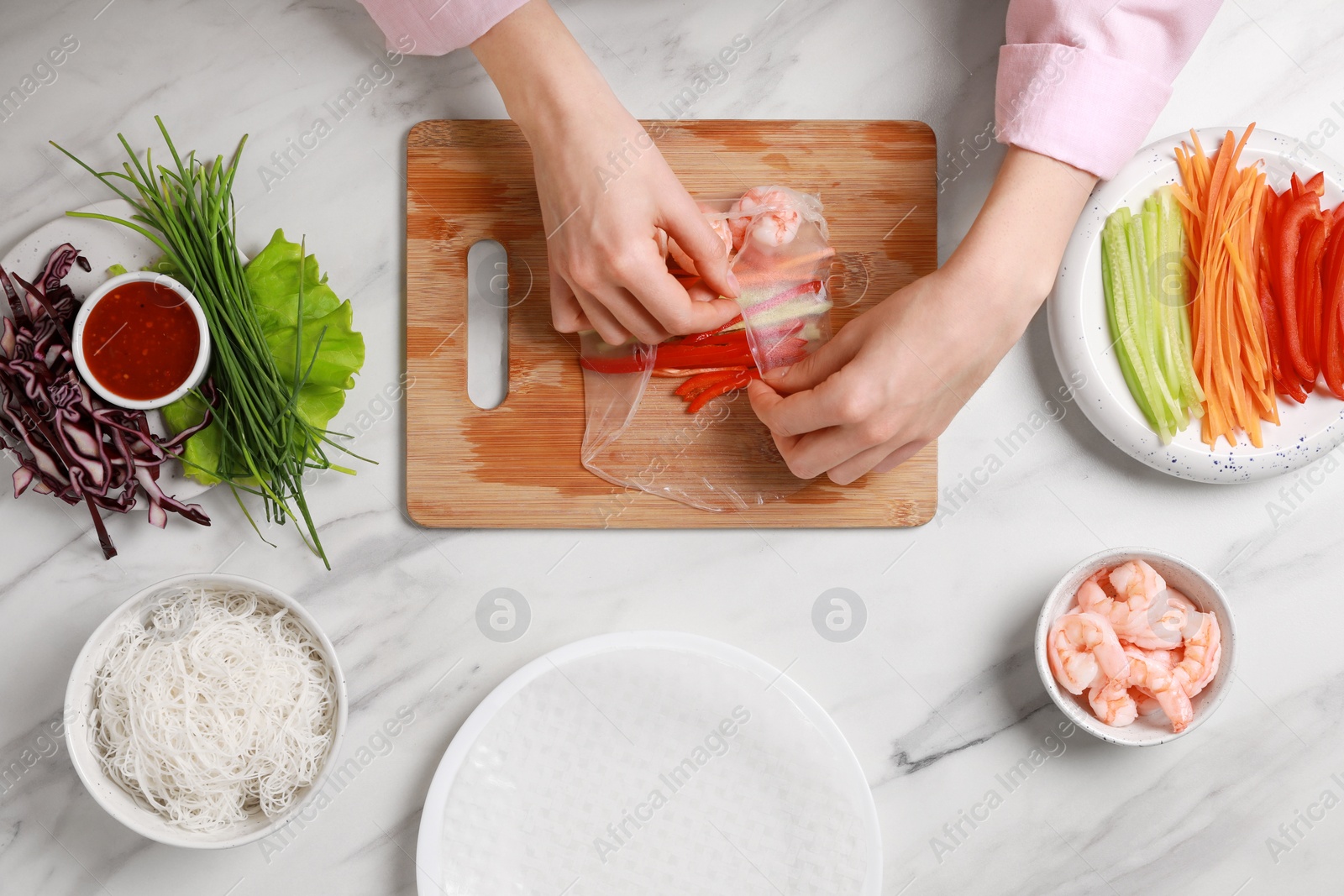 Photo of Making delicious spring rolls. Woman wrapping fresh vegetables and shrimps into rice paper at white marble table, top view