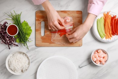 Photo of Making delicious spring rolls. Woman wrapping fresh vegetables and shrimps into rice paper at white marble table, top view