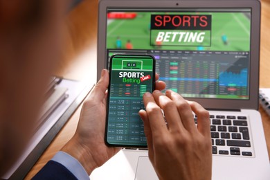 Image of Man betting on sports using smartphone and laptop at table, closeup. Bookmaker websites on displays