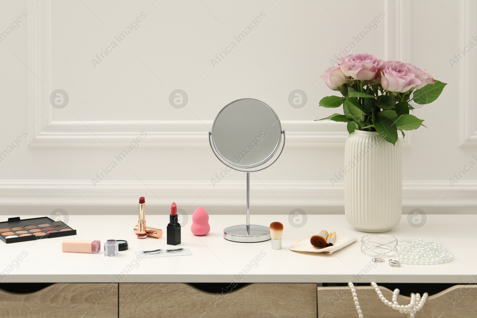 Photo of Mirror, cosmetic products, jewelry and vase with pink roses on white dressing table in makeup room