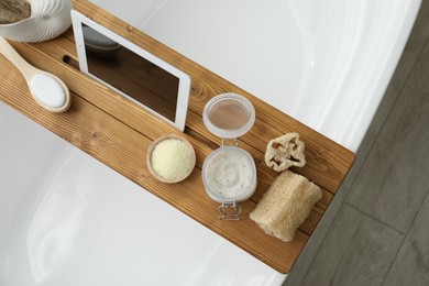 Wooden tray with tablet and spa products on bath tub in bathroom, top view