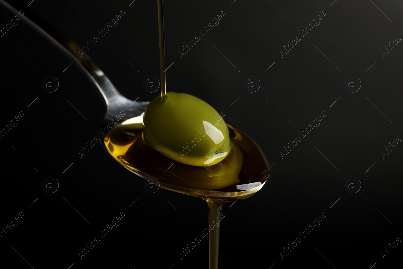 Photo of Pouring oil into spoon with ripe olive against black background, closeup