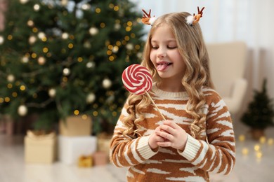 Cute girl with lollipop near Christmas tree at home. Space for text