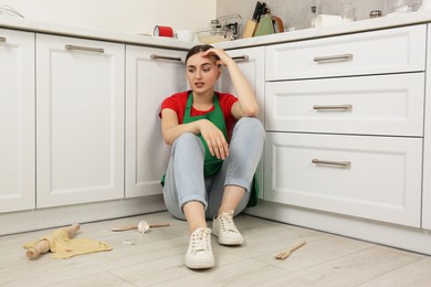 Photo of Stressed woman sitting on dirty floor with utensils and food leftovers in messy kitchen