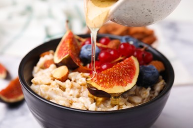 Pouring honey into bowl of oatmeal with berries, almonds and fig pieces on white table, closeup