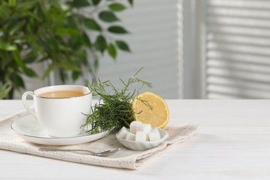 Photo of Aromatic herbal tea, fresh tarragon sprigs, sugar cubes and lemon on white wooden table, space for text