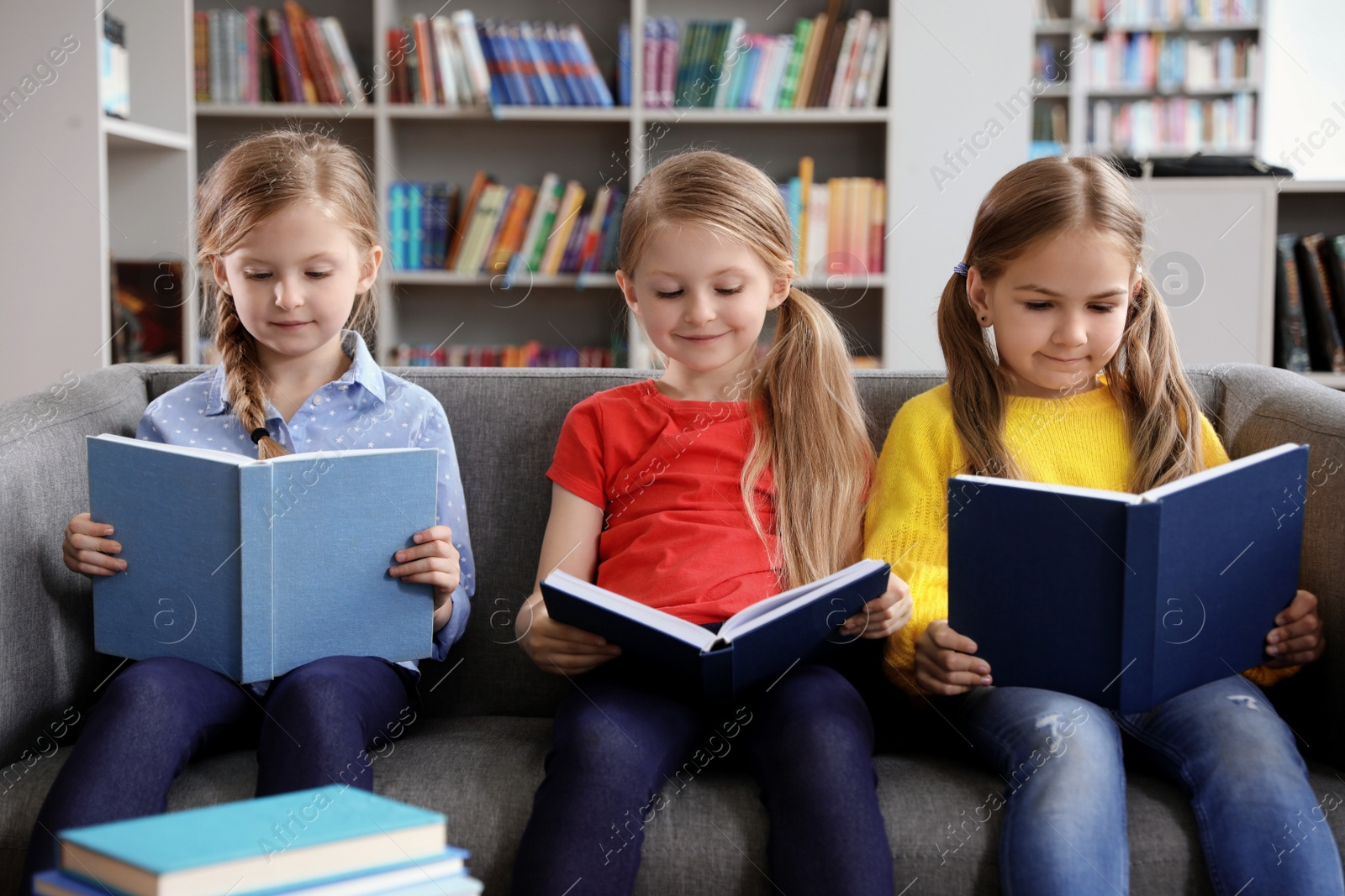 Photo of Happy little girls reading books on sofa in library