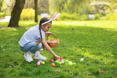 Photo of Easter celebration. Cute little girl in bunny ears hunting eggs outdoors, space for text