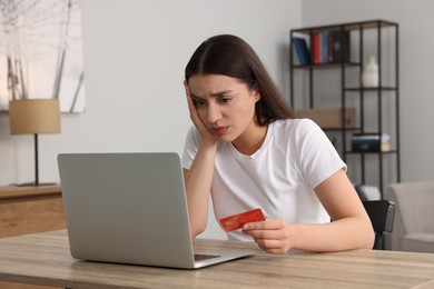 Photo of Upset woman with credit card using laptop at table indoors. Be careful - fraud